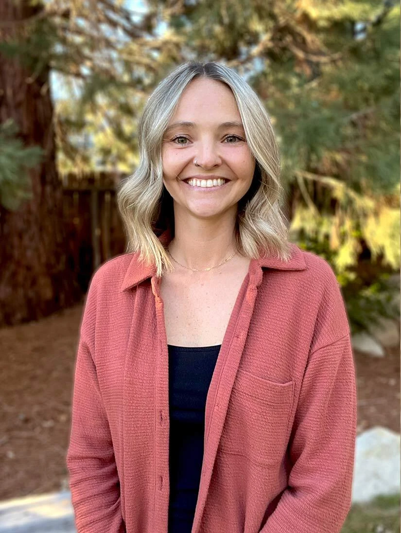 A woman with blonde hair and a light-colored sweater posing in front of trees