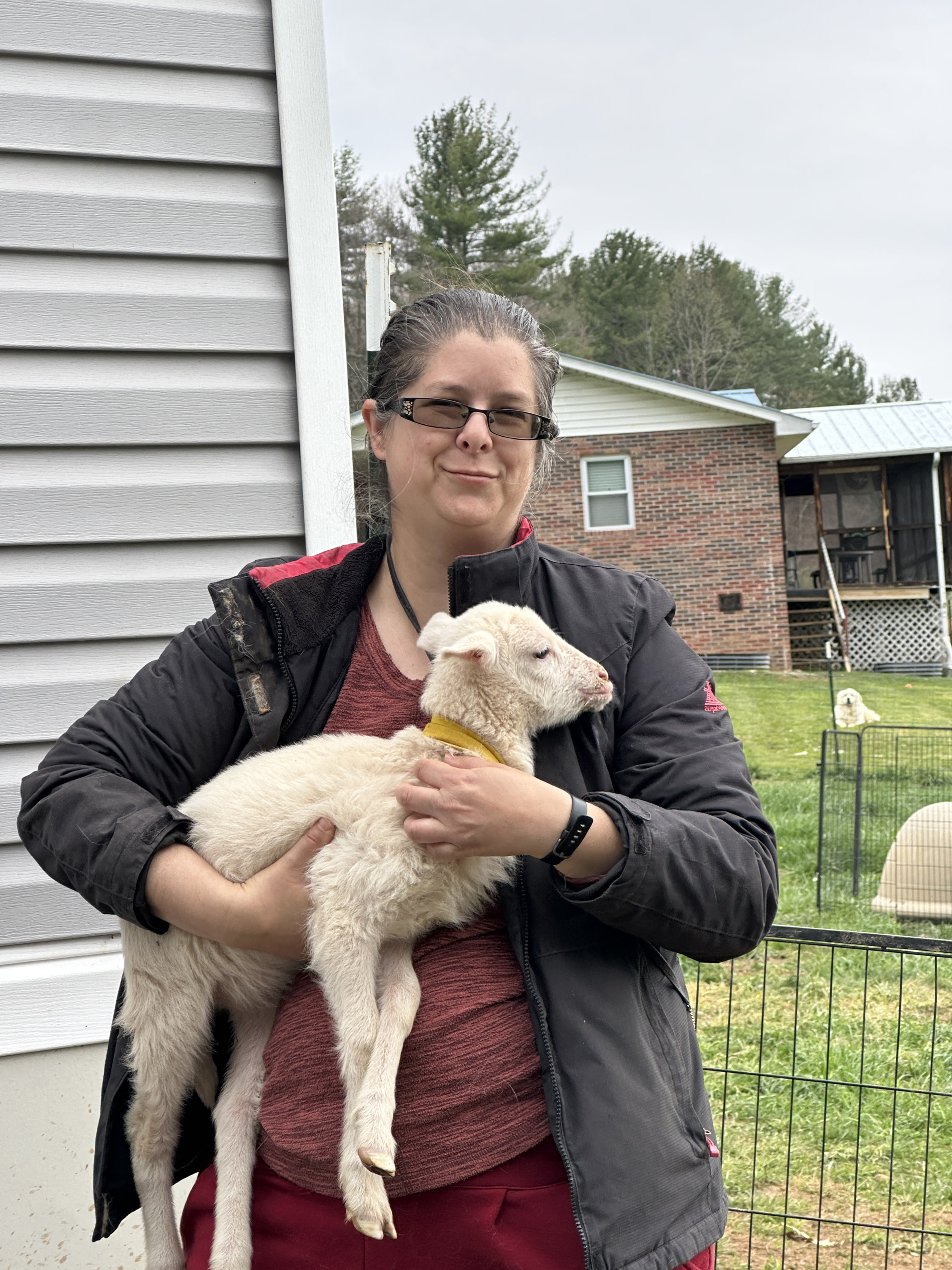 A woman wearing red clothes underneath a dark coat holding a young sheep while facing the photographer